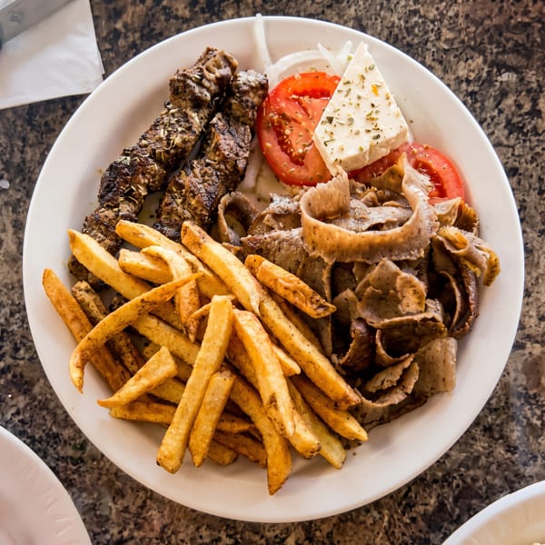 A white plate with grilled meat skewers, gyro meat, french fries, tomatoes, onions, and feta cheese.