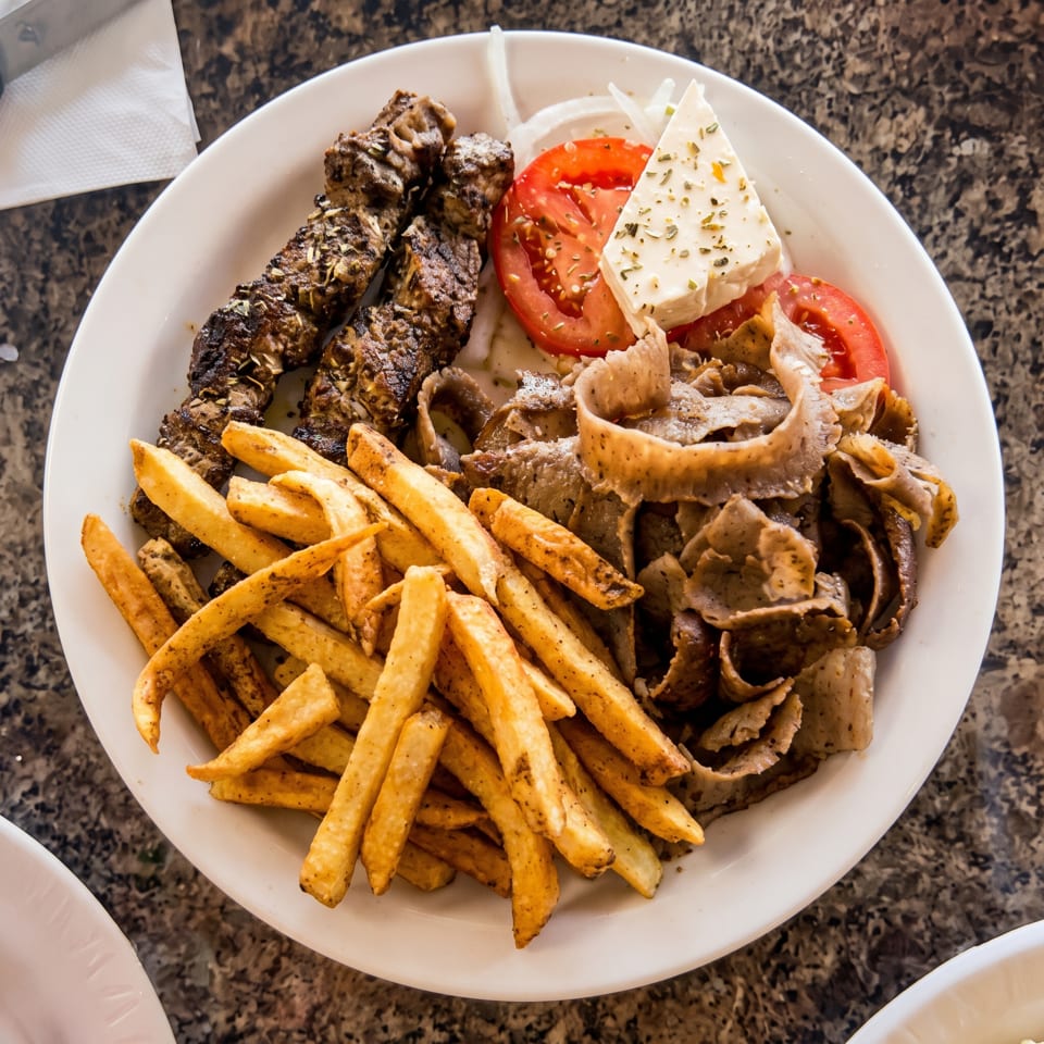 A white plate with grilled meat skewers, gyro meat, french fries, tomatoes, onions, and feta cheese.