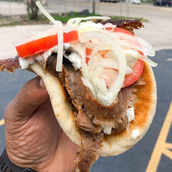 Hand holding a gyro with meat, tomatoes, onions, and white sauce, outdoors with a blurred background.