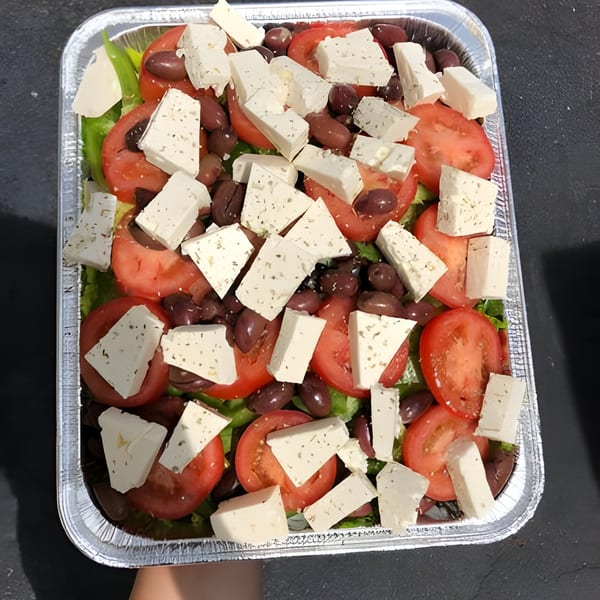 A large Greek salad with tomatoes, olives, and feta cheese in a foil pan.