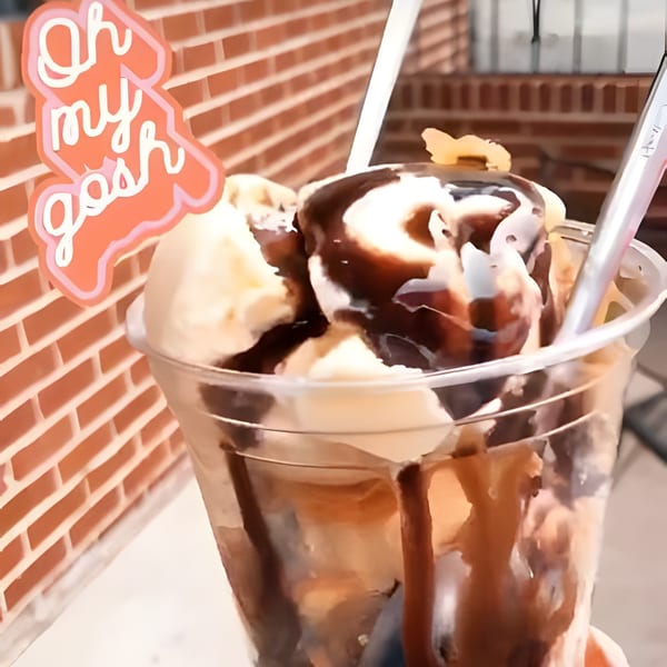 A clear plastic cup filled with vanilla ice cream, chocolate syrup, and a spoon, against a brick wall.