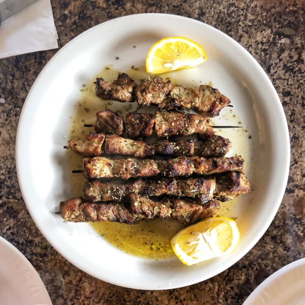 Grilled pork souvlaki skewers with lemon wedges and olive oil on a white plate.